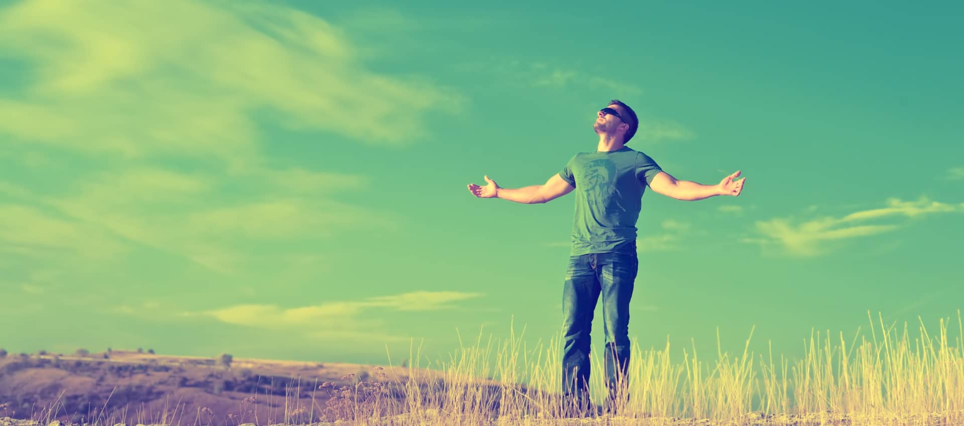 white man in dead grass file,d blind folded arms up head tilted towards sky, sky behind him